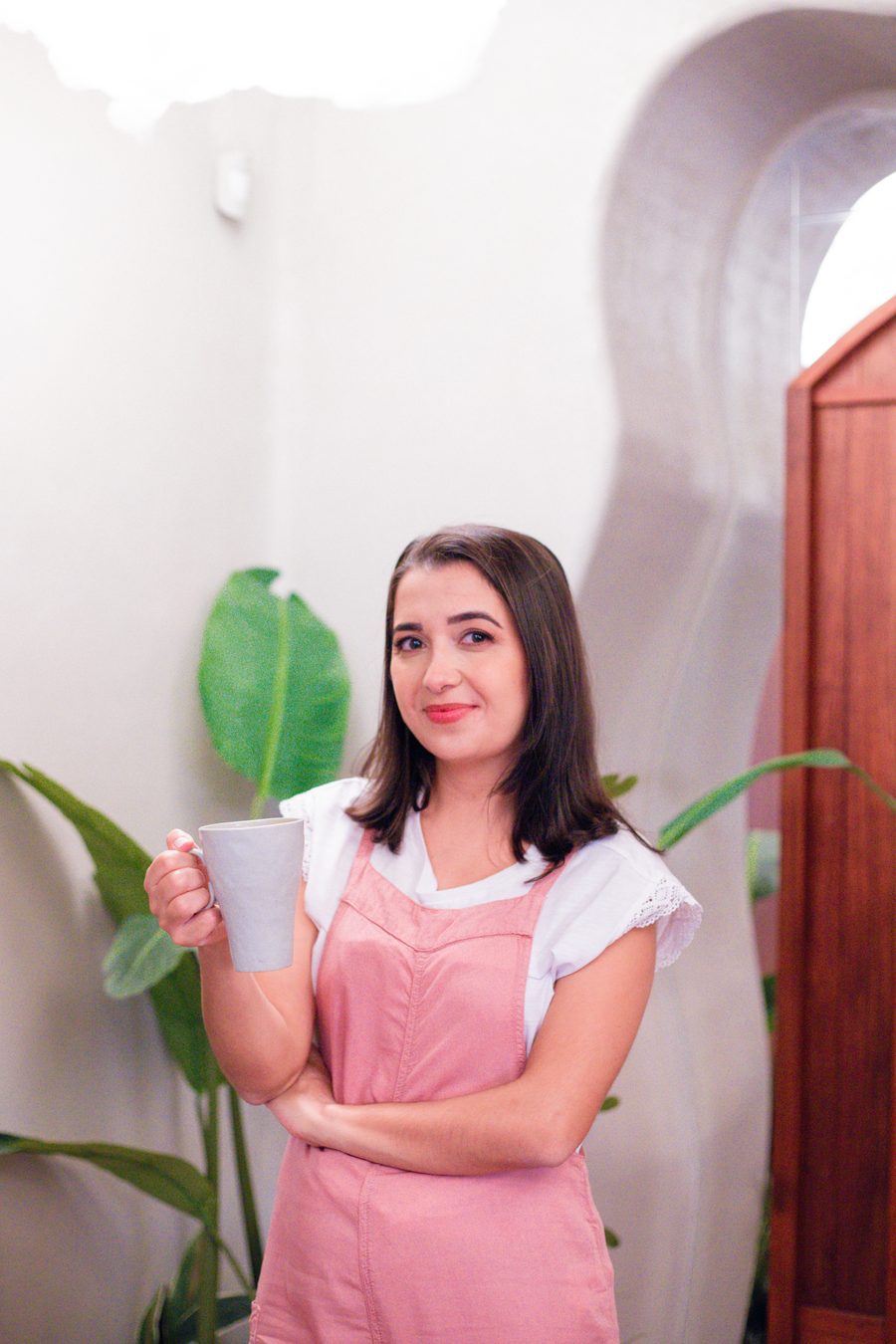Grazielle Bonfim sorrindo, com uma xícara na mão, de camiseta branca e jardineira rosa, ao lado de plantas