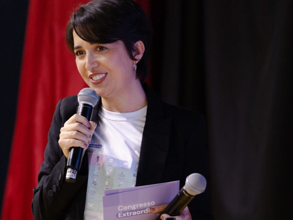 Grazielle sorrindo no palco, com um microfone na mão, em um congresso